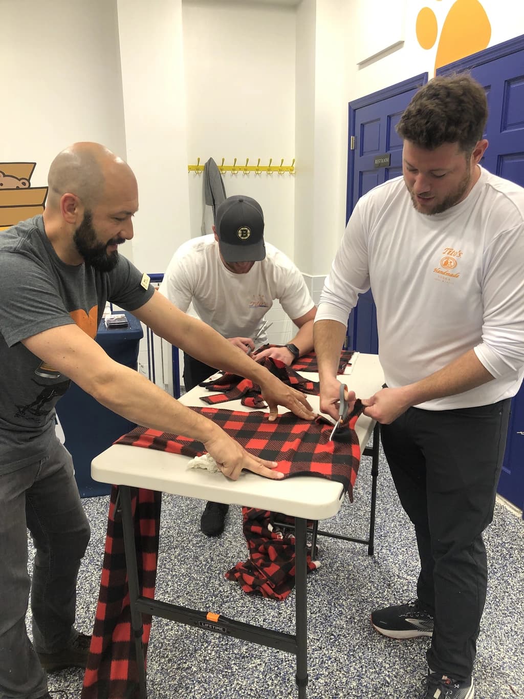 Three men cut red and black plaid fabric on a table, concentrating on creating blankets in a collaborative crafting activity.