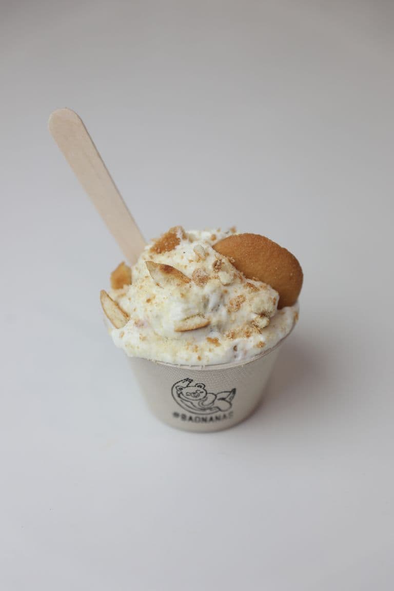 Single serving of banana pudding in a branded cup, topped with crumbled cookies and banana slices, with a wooden spoon inserted.