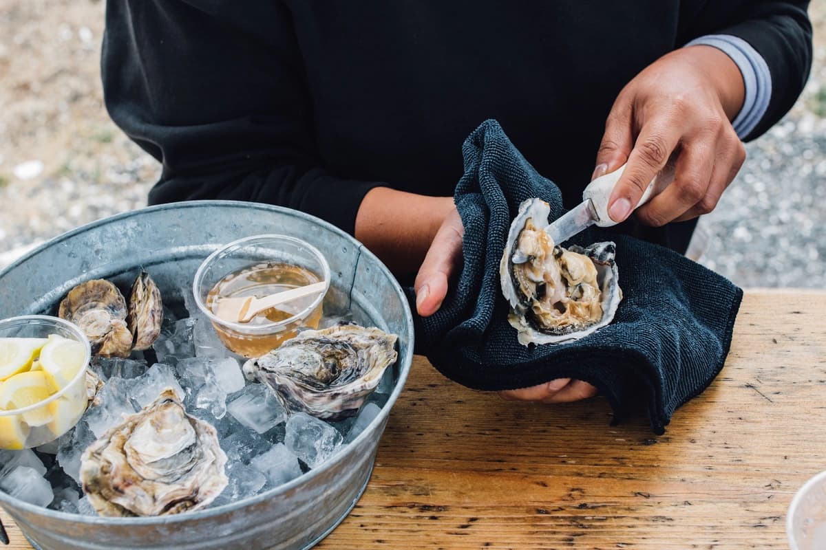 Hands shucking an oyster over a bucket of ice containing more oysters and lemon wedges, with a cup of sauce nearby.
