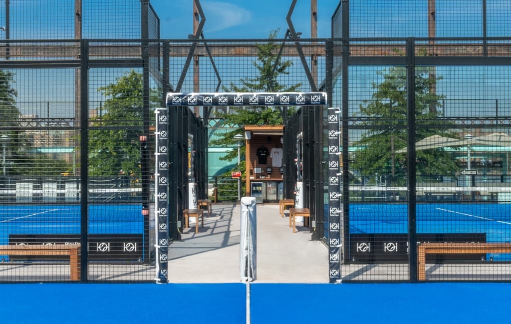 Empty padel court with blue flooring and glass walls, ready for players, under a clear blue sky.