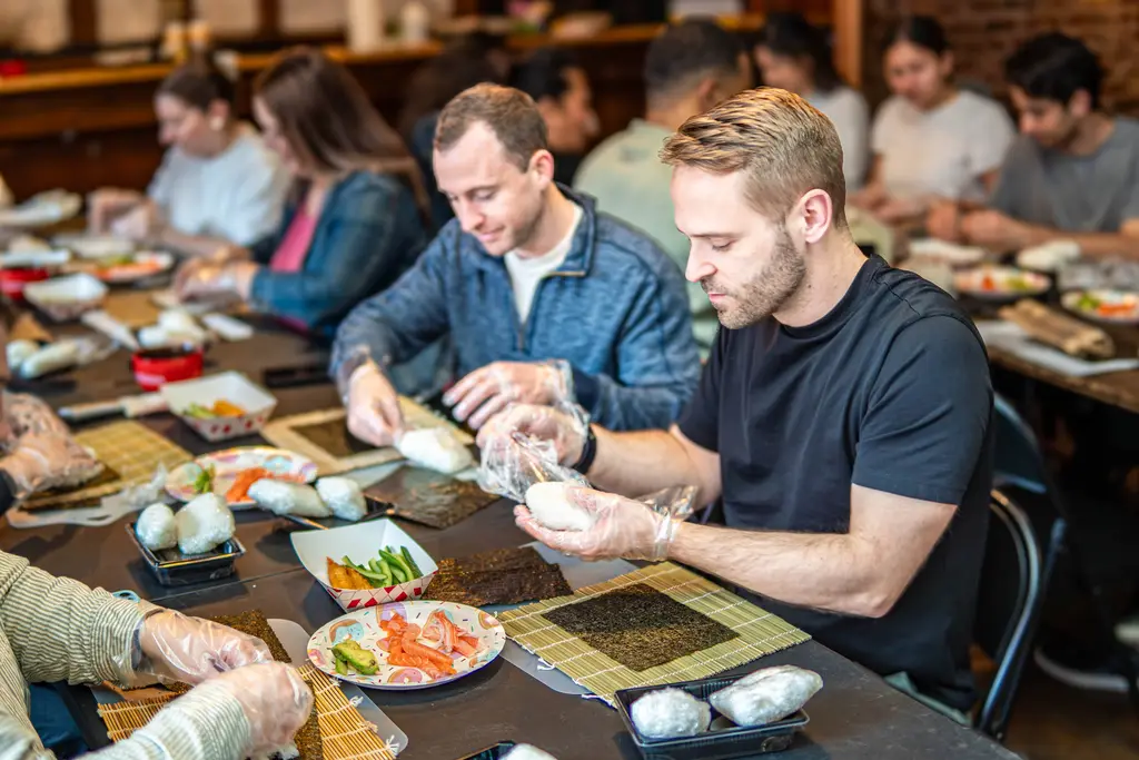 A group of people intensely focused on making sushi, carefully wrapping rolls in seaweed and rice, all seated around a long communal table.