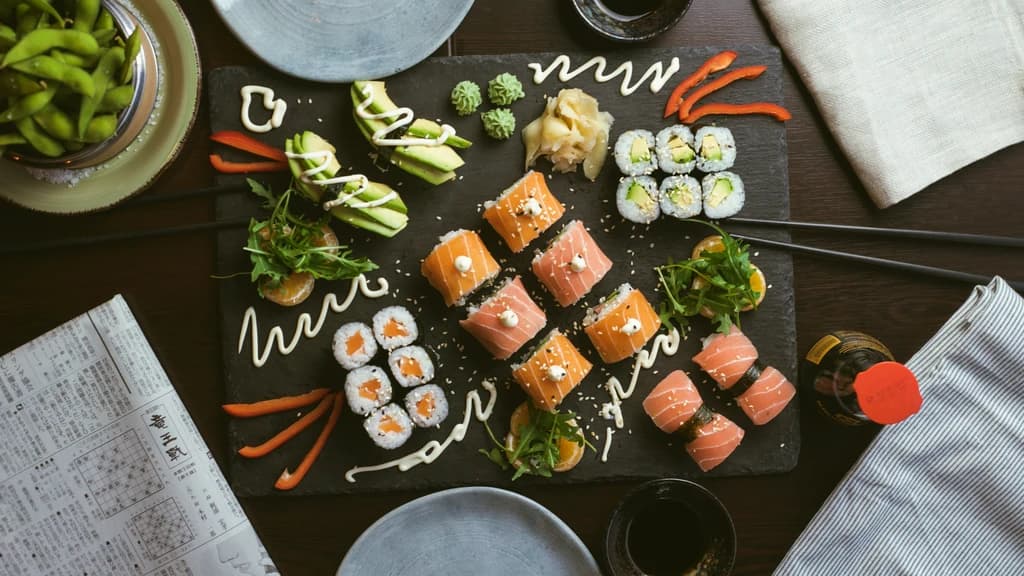A beautifully arranged sushi platter on a dark slate board, featuring various rolls, fresh slices of fish, and garnishes, with chopsticks and side dishes on a wooden table.
