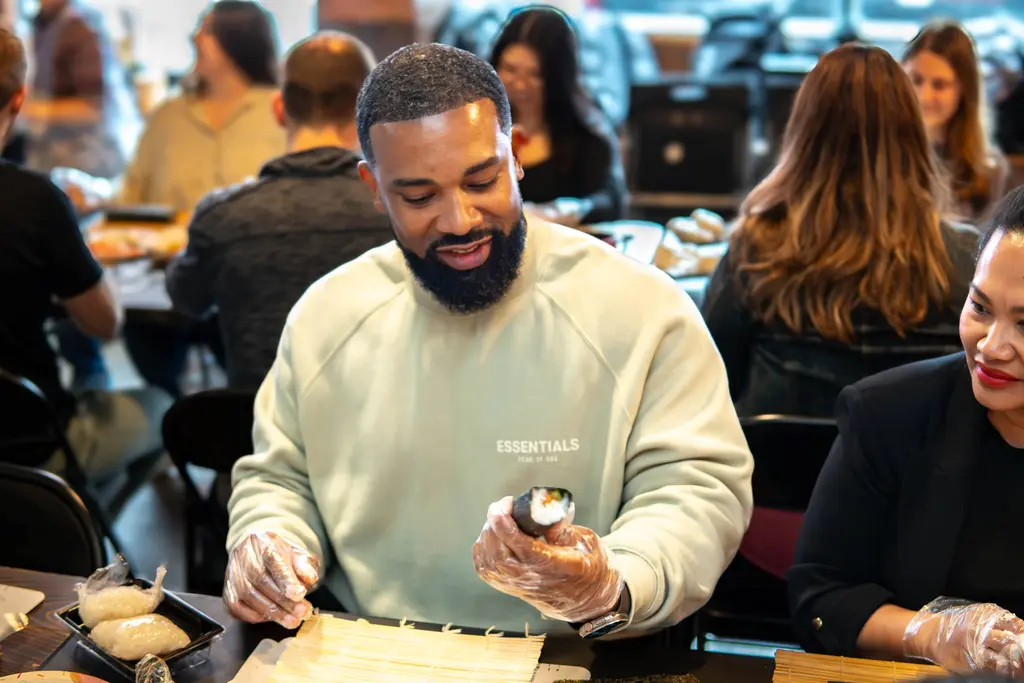 A man smiling while holding a sushi roll he made during a hands-on cooking class, wearing gloves and seated at a table with other participants.