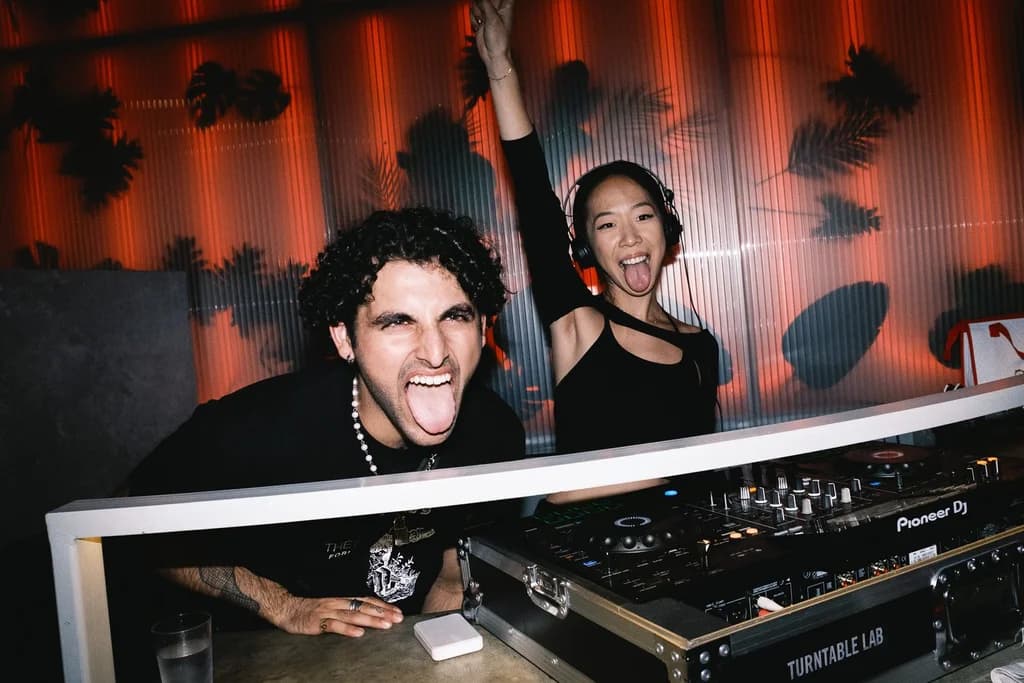 Two DJs behind a booth, smiling and making playful expressions with vibrant lighting in the background.