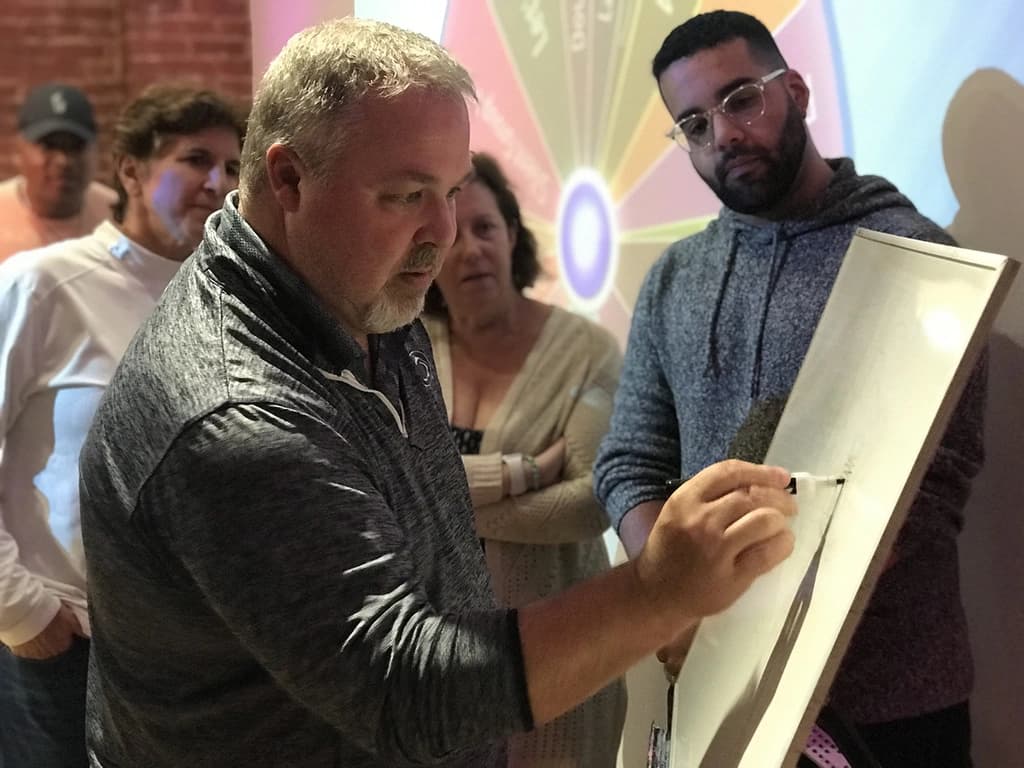 A man focused on drawing on a large notepad, while a group of people watches attentively during a team-building exercise.