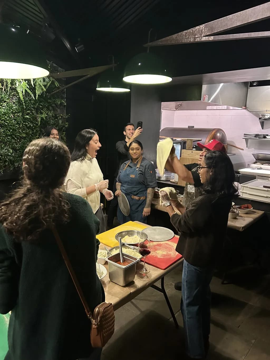 A group of people gathered around a table, stretching pizza dough and preparing pizzas, while a chef supervises.