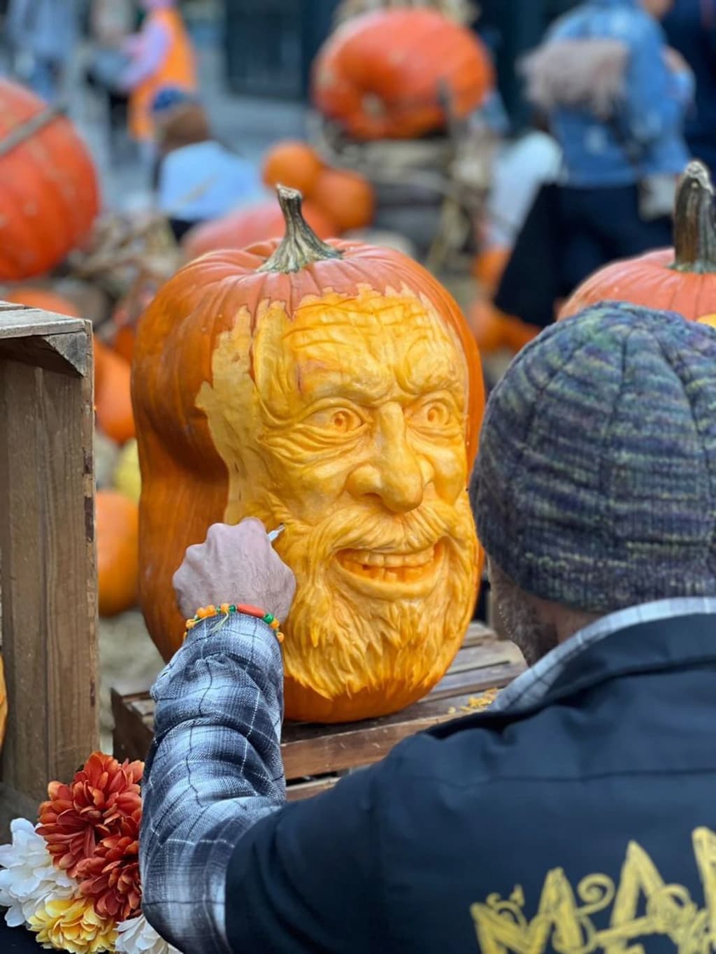 An artist meticulously carves a pumpkin into a detailed face at a pumpkin carving demonstration, surrounded by other pumpkins.