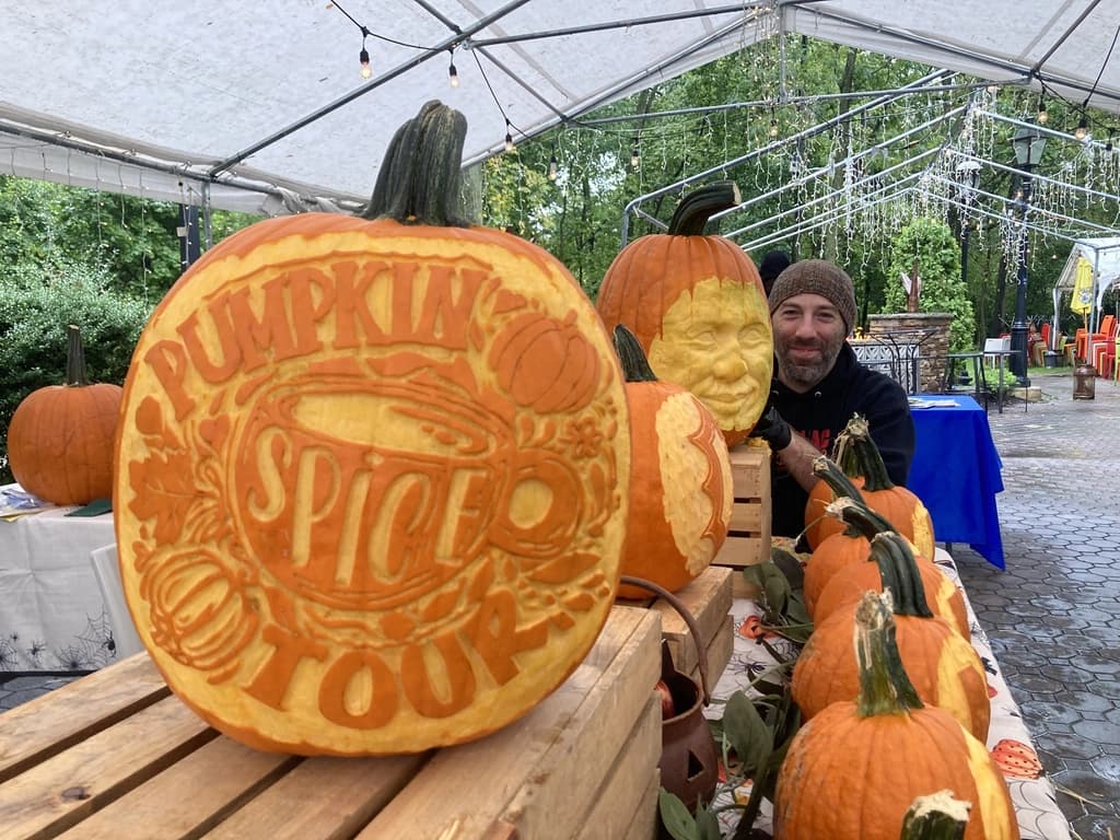 Pumpkin intricately carved with the words 'Pumpkin Spice Tour.' Artist and other carved pumpkins are in the background under a tent.