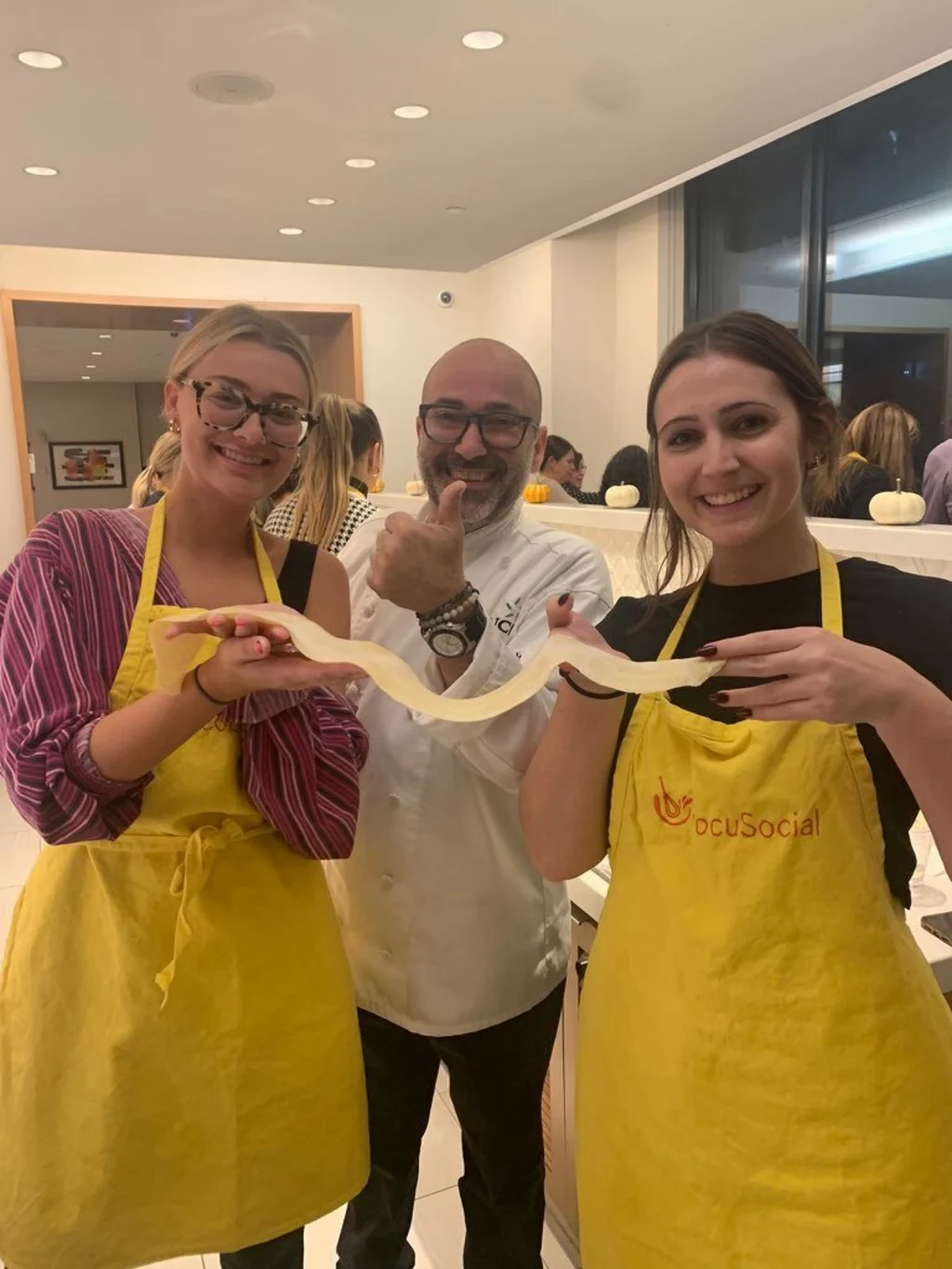 Two women and a chef in yellow aprons proudly display a long strip of freshly made pasta dough, smiling at the camera.