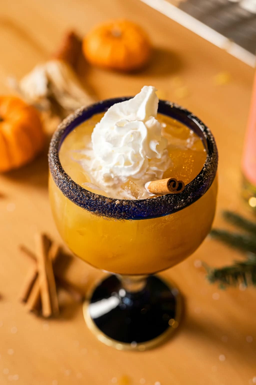 A large margarita glass filled with a pumpkin-colored drink, topped with whipped cream and a cinnamon stick garnish.