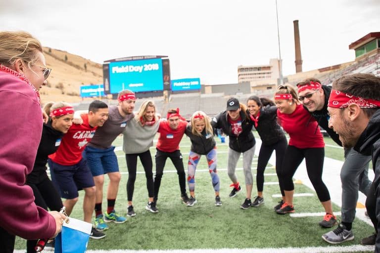 Team in red bandanas huddles on a field, smiling and getting hyped with “Field Day 2018” on the stadium screen behind them.