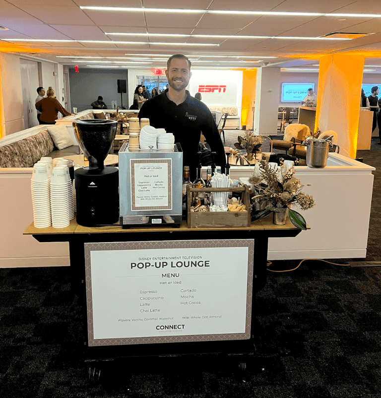 A smiling barista stands behind a pop-up coffee cart in an ESPN office, offering espresso drinks from a menu labeled "Disney Entertainment Television Pop-Up Lounge.