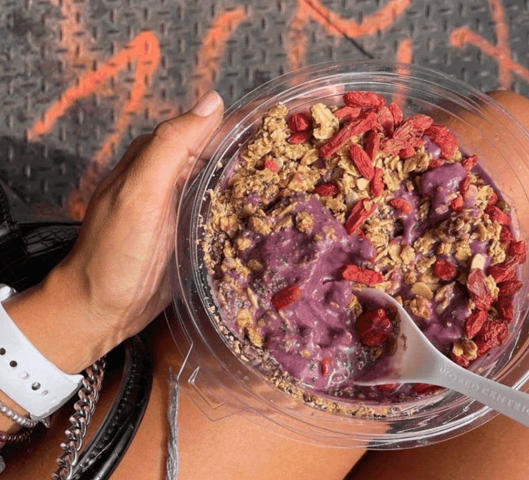 Açaí bowl topped with granola and goji berries, served in a clear container with a spoon, held by a person sitting outdoors.
