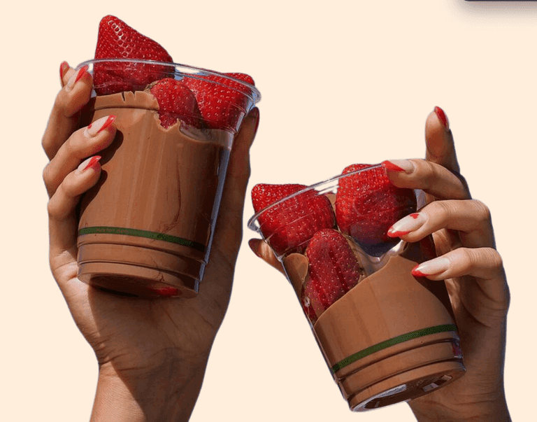 Two hands holding clear cups filled with fresh strawberries and creamy chocolate, with red and white manicured nails complementing the treat.