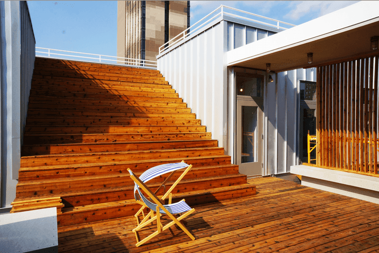 "A modern rooftop terrace featuring wide wooden steps that double as seating, bathed in warm sunlight. A blue and white striped wooden deck chair sits on the wooden deck floor. The space includes a white container-style structure with glass doors and wooden slatted railings. White safety railings line the upper portion of the terrace. A tall brick building is visible in the background against a clear blue sky. The architecture combines industrial elements with warm wood tones, creating a contemporary urban outdoor space."RetryClaude can make mistakes. Please double-check responses.