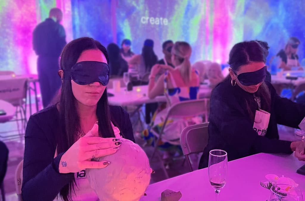 Two women wearing blindfolds, engaged in a creative activity, sculpting clay heads while seated in a vibrant room with colorful lights.
