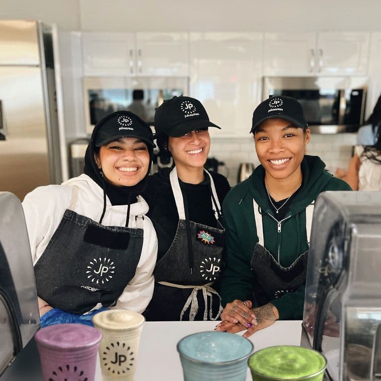 Three smiling Juice Press employees in branded aprons and hats stand behind a counter with colorful cups in a bright kitchen setting.