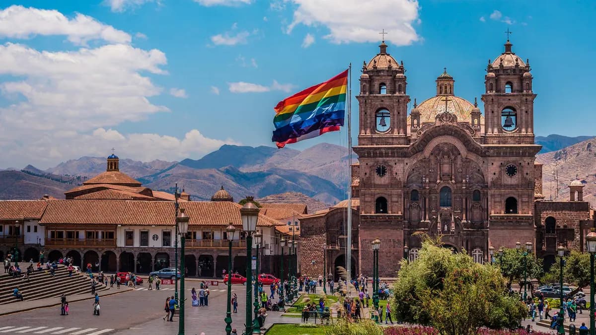 A vibrant plaza in Cusco, Peru, featuring a cathedral, a rainbow flag, and people enjoying the sunny day.