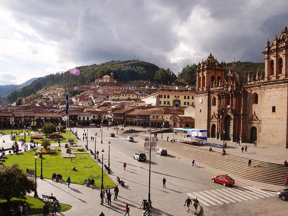 A bustling plaza in Cusco, Peru, with colonial architecture, green lawns, and people walking around.