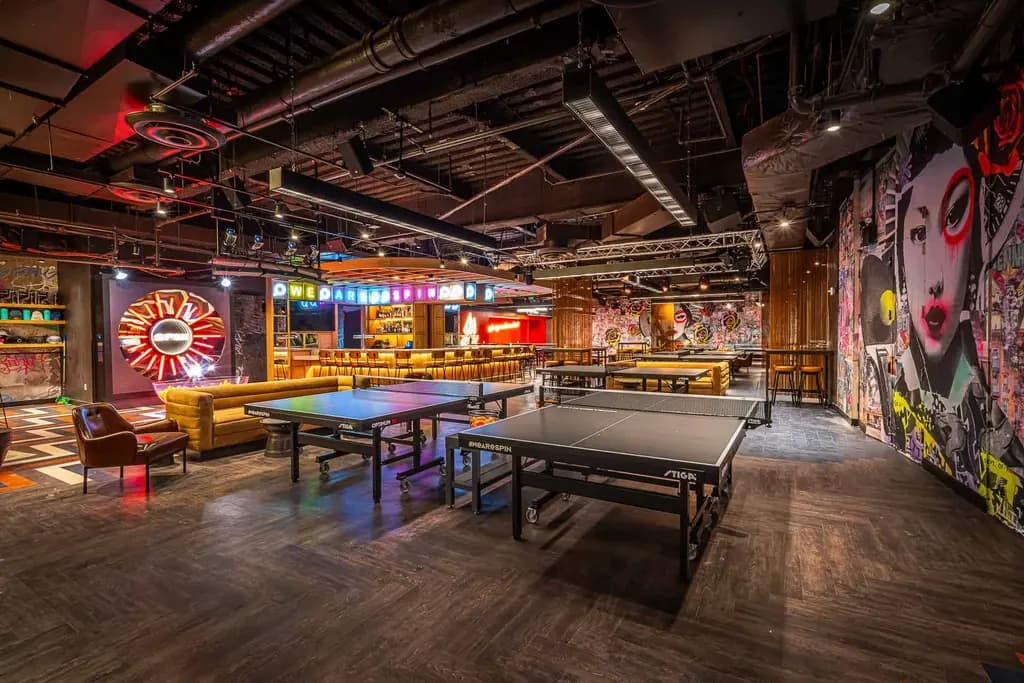 Vibrant indoor venue with colorful graffiti walls, neon lights, and multiple ping pong tables, creating an energetic and fun environment.
