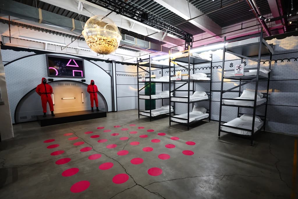 Squid Game-inspired room with pink guards and bunk beds. A giant cash-filled orb hangs from the ceiling, evoking the show's ominous tone.