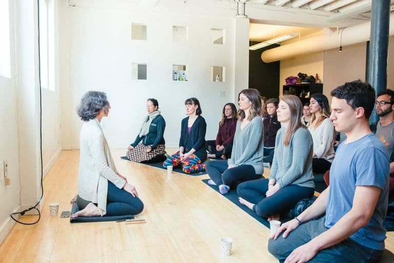 Group meditation session with a guide leading participants seated on mats in a bright studio.