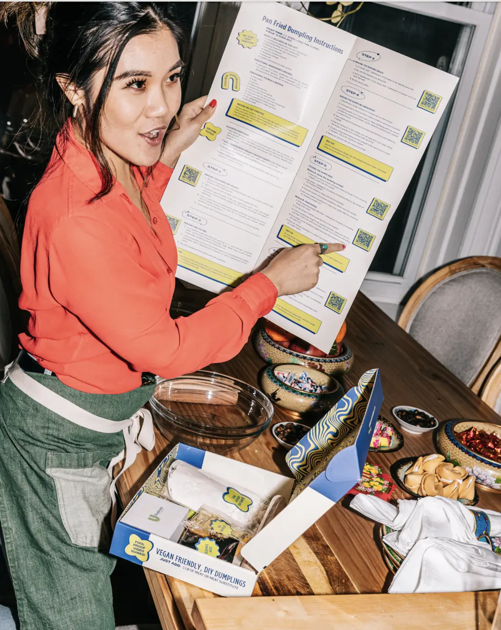 A person in a red shirt holding a step-by-step guide for making dumplings, with a DIY dumpling kit and colorful bowls on the table.