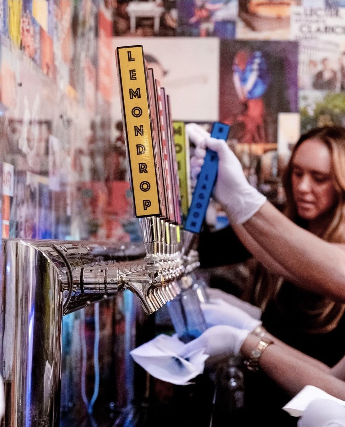 Close-up of a person filling bottles with colorful liquids from taps labeled with different flavors in a tie-dye workshop.
