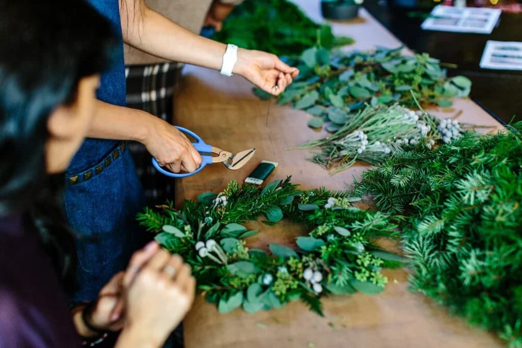 A person using blue-handled scissors to cut greenery, preparing to create a holiday wreath. Several types of greenery and other materials are spread out on the table.
